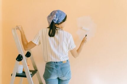 A woman with a bandana using a paint roller to paint a peach-colored wall indoors.