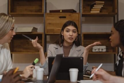 Three women engaged in a collaborative meeting with laptops and notes in an office setting.